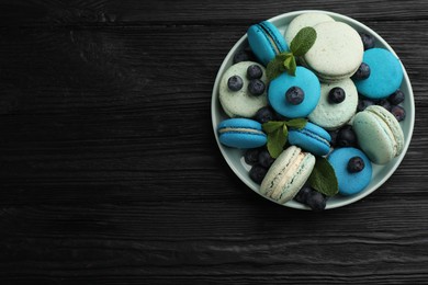 Delicious macarons, mint and blueberries in bowl on black wooden table, top view. Space for text