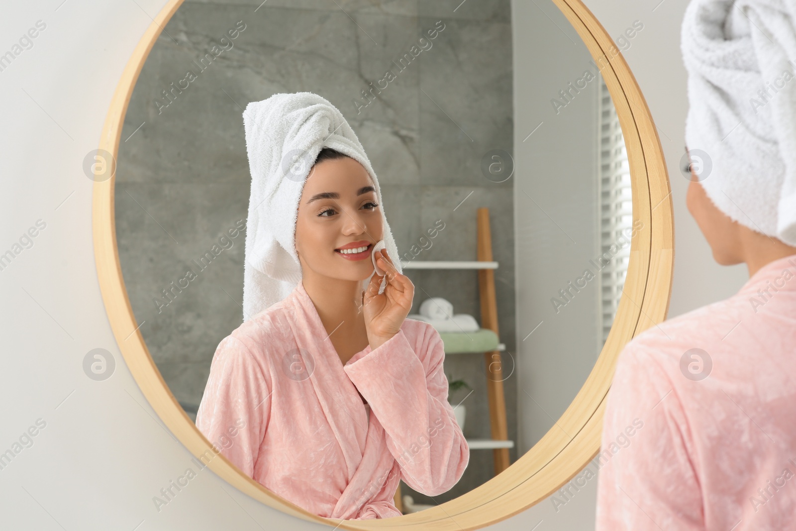 Photo of Beautiful woman in terry towel removing makeup with cotton pad near mirror indoors