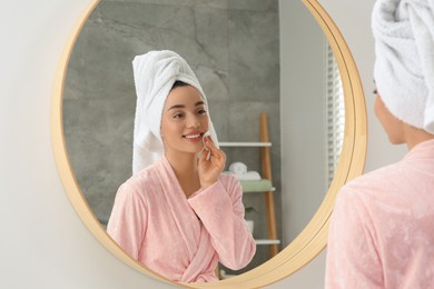 Photo of Beautiful woman in terry towel removing makeup with cotton pad near mirror indoors