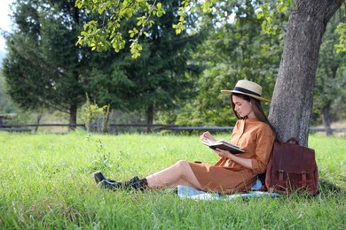 Beautiful young woman reading book near tree on green grass