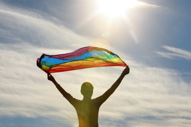 Woman holding bright LGBT flag against blue sky on sunny day, back view