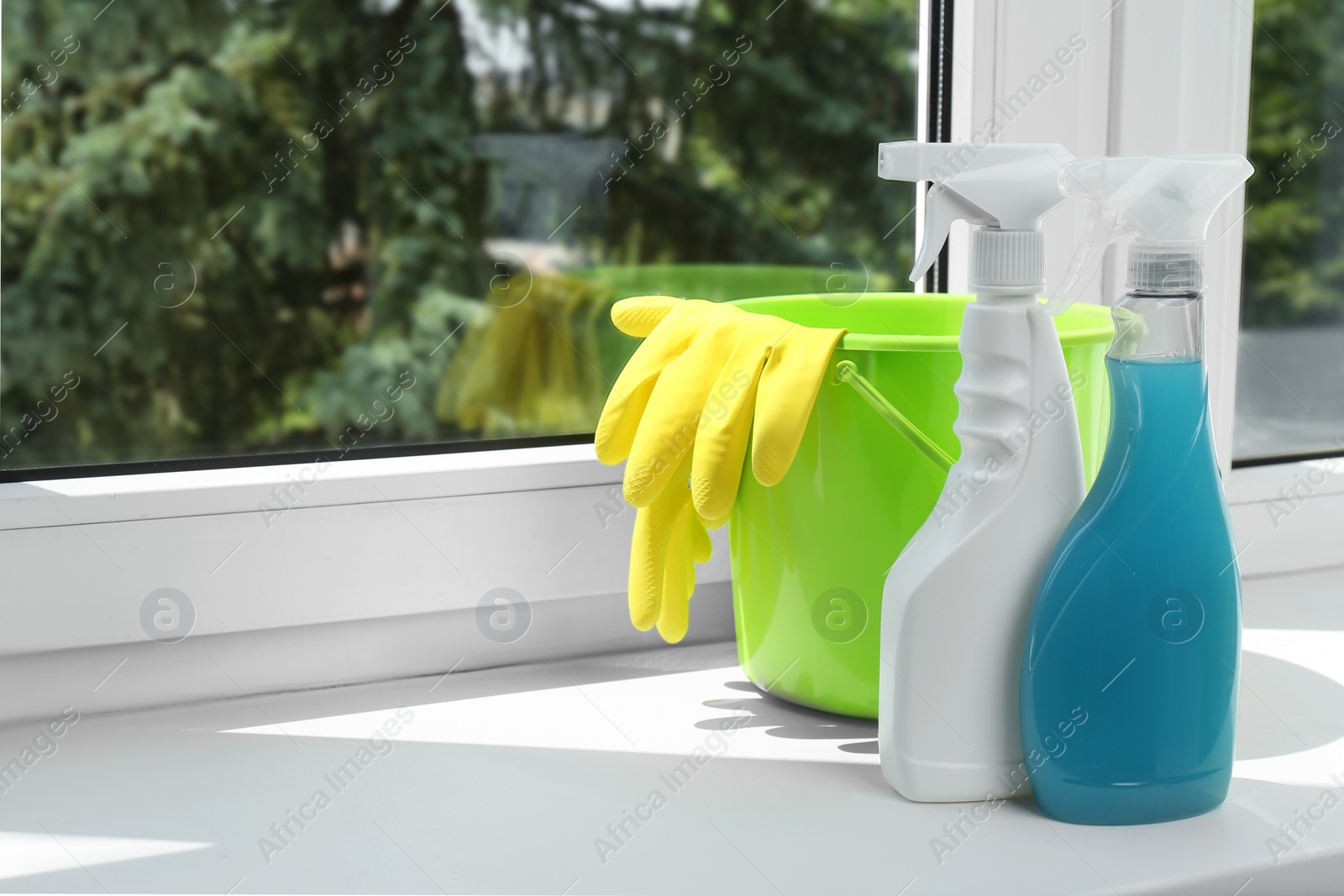 Photo of Green bucket with gloves and spray bottles of detergents on window sill indoors, space for text. Cleaning supplies