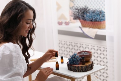 Beautiful young woman with bottle of essential oil at table indoors