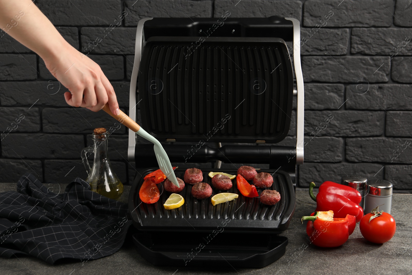 Photo of Woman cooking meat balls with bell peppers and lemon on electric grill at grey textured table, closeup