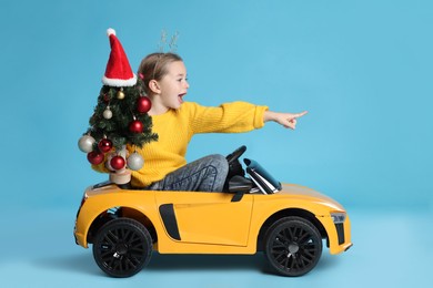 Photo of Cute little girl with Christmas tree driving children's electric toy car on light blue background