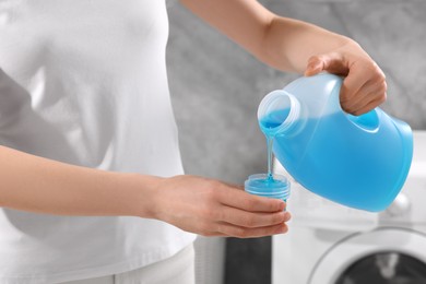 Photo of Woman pouring fabric softener from bottle into cap for washing clothes indoors, closeup