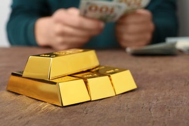 Stacked gold bars and man counting money at table, closeup. Space for text