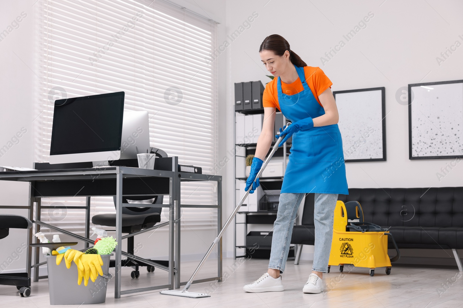 Photo of Cleaning service worker washing floor with mop. Bucket with supplies and wet floor sign in office