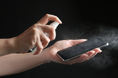 Photo of Woman spraying antiseptic onto smartphone against black background, closeup