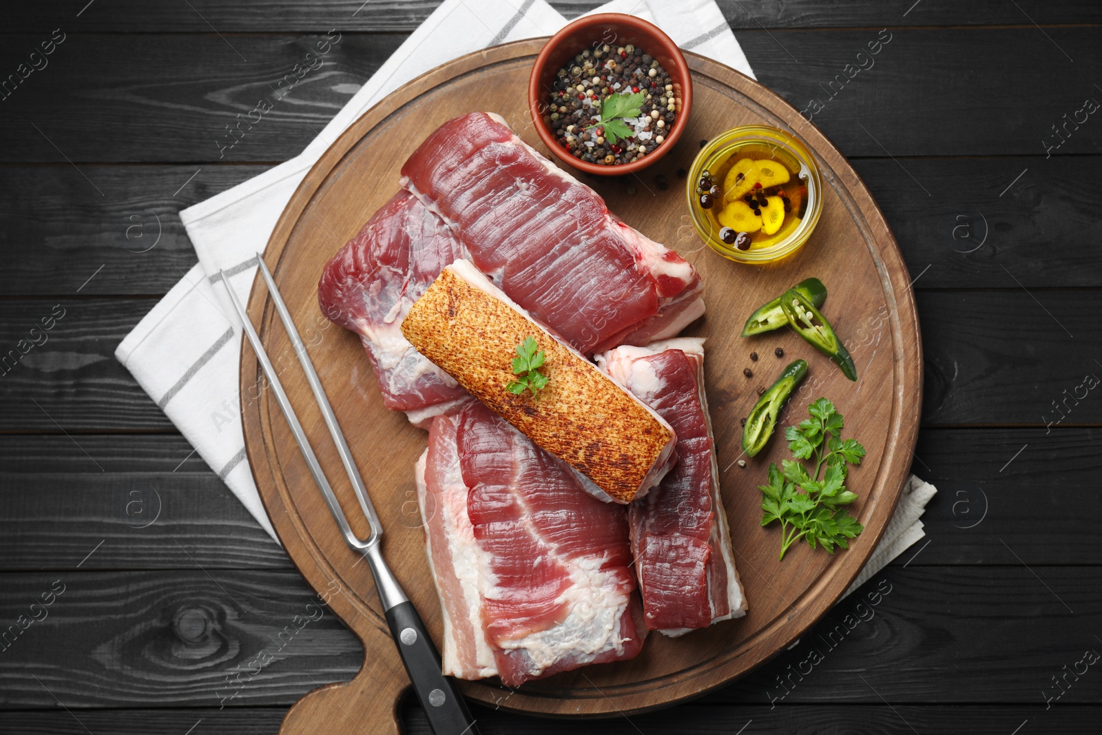 Photo of Pieces of raw pork belly, chili pepper, peppercorns, oil and parsley on black wooden table, top view