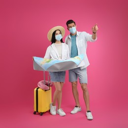 Photo of Couple of tourists in medical masks with map and suitcase on pink background. Travelling during coronavirus pandemic