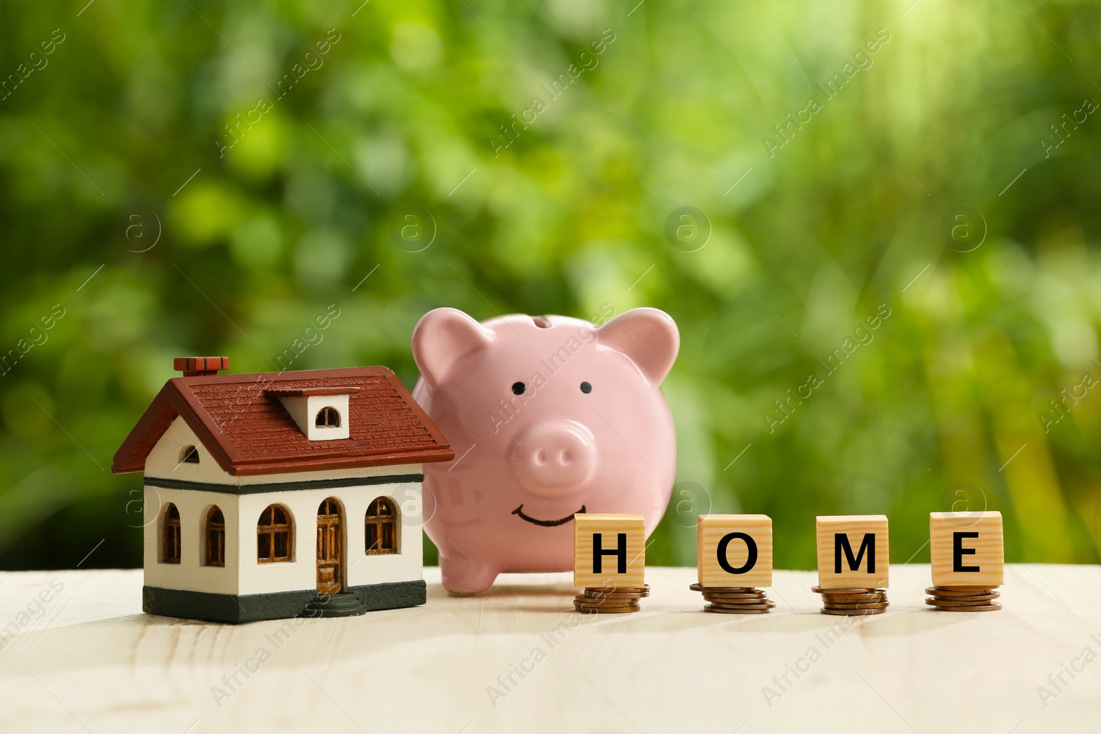 Photo of Piggy bank, word Home made of cubes, house model and stacks of coins on wooden table outdoors