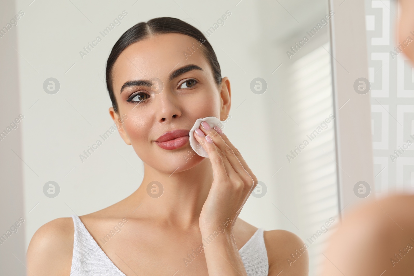 Photo of Beautiful woman removing makeup with cotton pad near mirror indoors