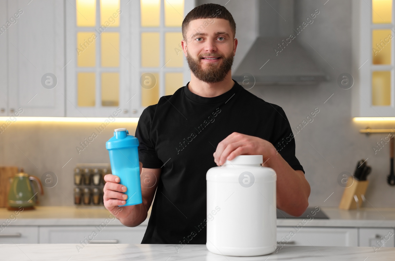 Photo of Young man with shaker of protein and powder at white marble table in kitchen