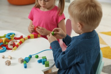 Little children playing with wooden pieces and string for threading activity at white table indoors. Developmental toys