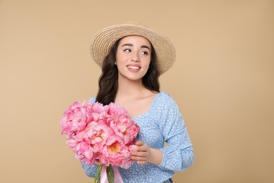 Beautiful young woman in straw hat with bouquet of pink peonies against beige background