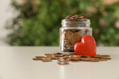 Photo of Red heart and donation jar with coins on table against blurred background. Space for text