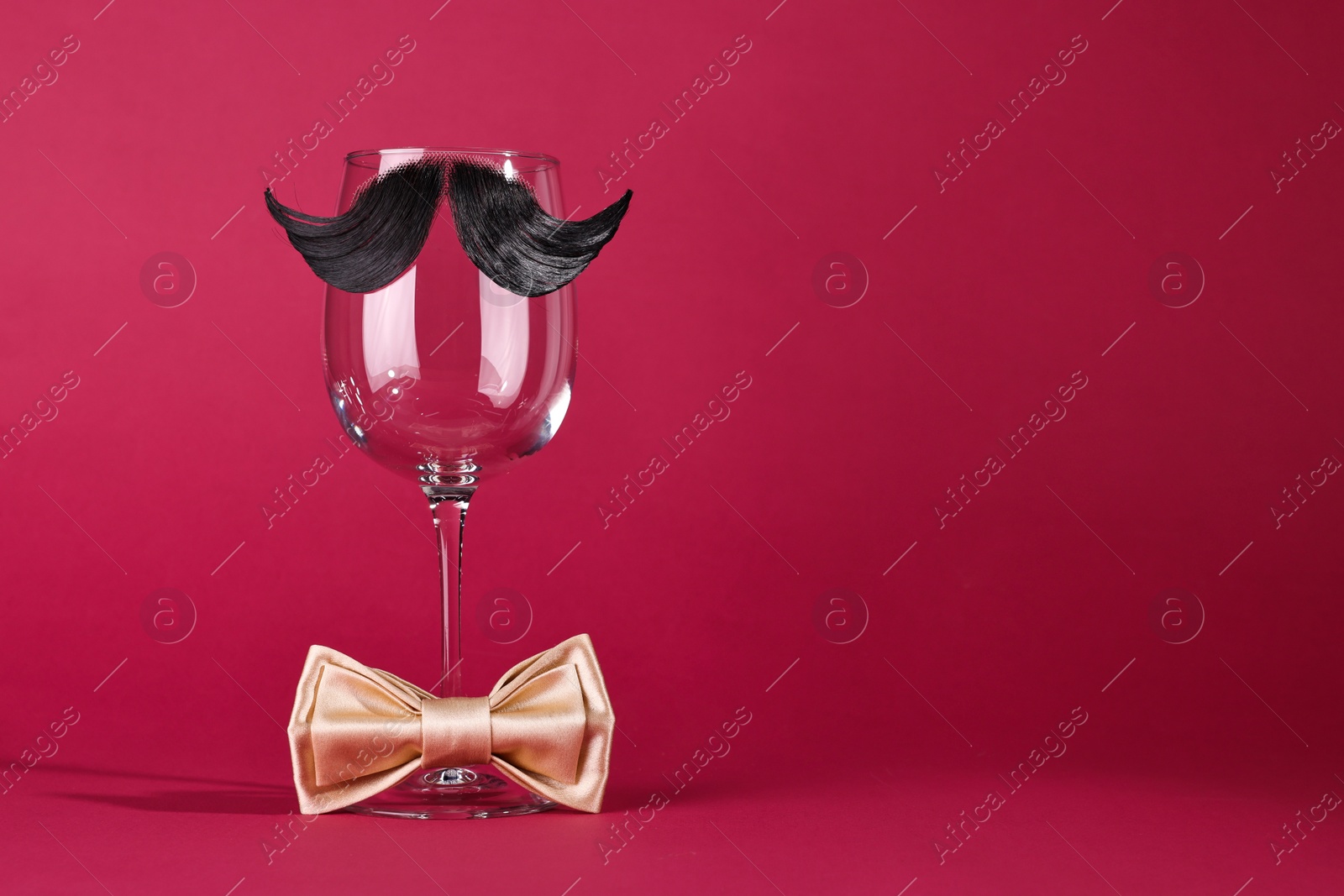 Photo of Man's face made of artificial mustache, bow tie and wine glass on crimson background. Space for text