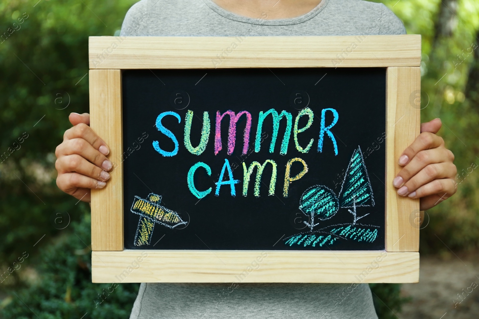 Photo of Woman holding blackboard with text SUMMER CAMP and drawing on blurred background, closeup