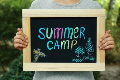 Photo of Woman holding blackboard with text SUMMER CAMP and drawing on blurred background, closeup