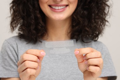 Photo of Young woman holding teeth whitening strips on light grey background, closeup