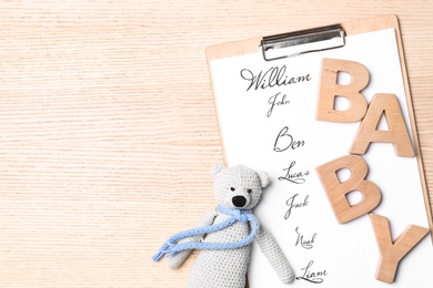 Photo of Clipboard with written baby names, wooden letters and toy on wooden table, flat lay. Space for text