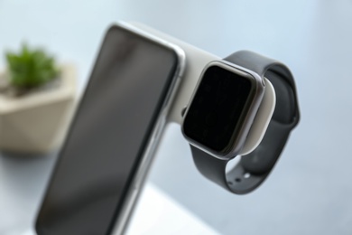 Photo of Stylish smart watch and phone charging with wireless pad against blurred background, closeup