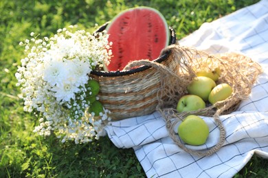 Photo of Picnic blanket with tasty fruits, beautiful flowers and basket on green grass outdoors