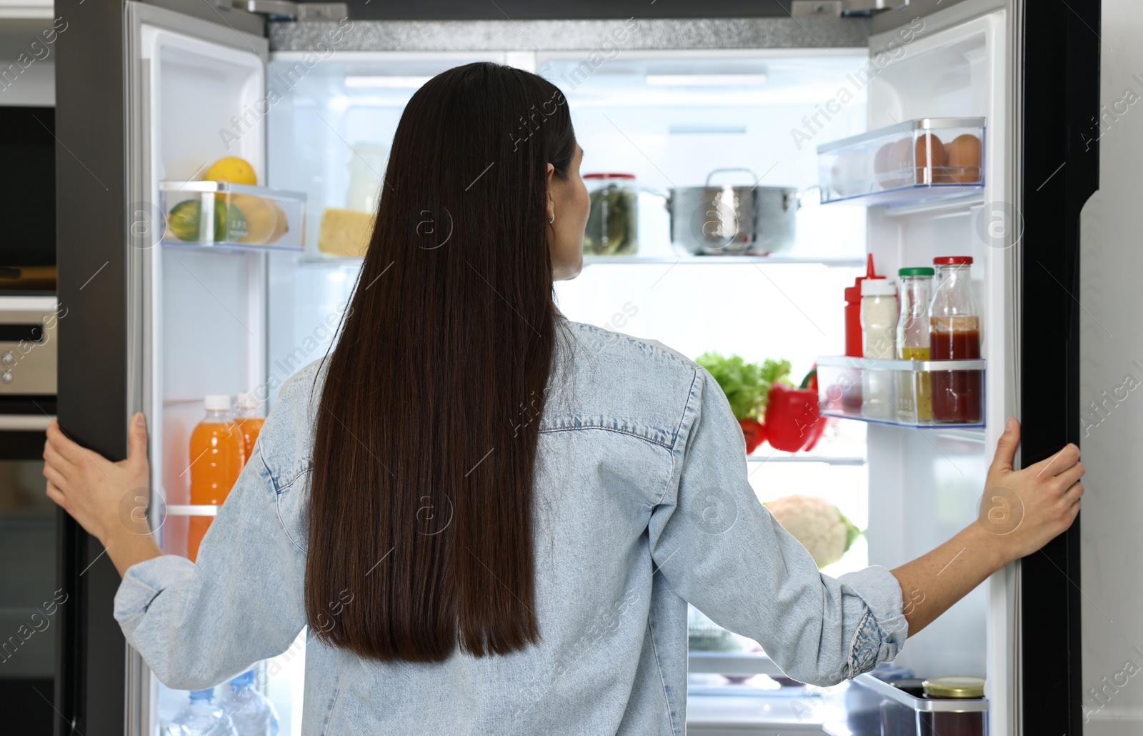 Photo of Young woman near modern refrigerator in kitchen, back view