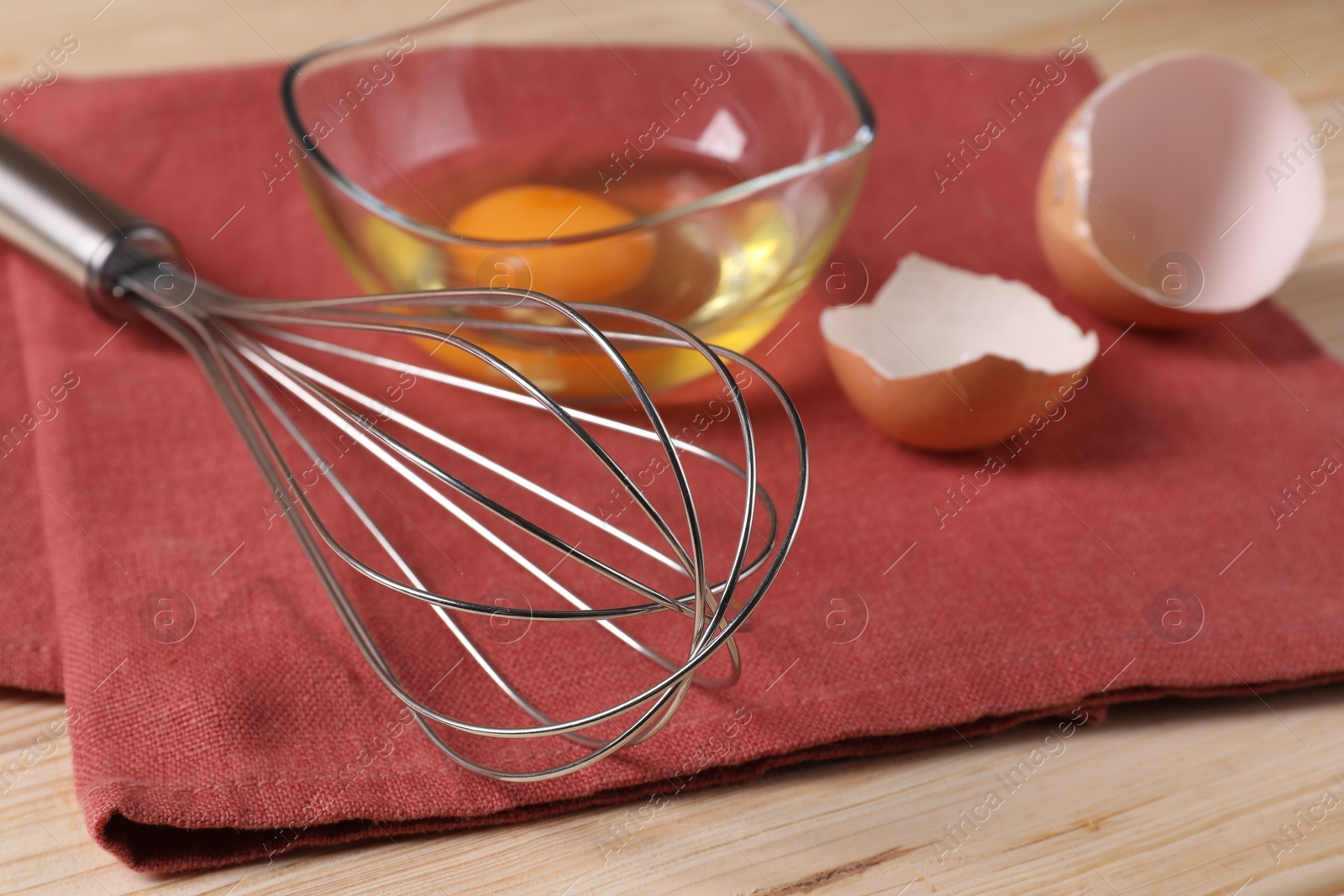 Photo of Metal whisk, raw eggs in bowl and shells on wooden table, closeup