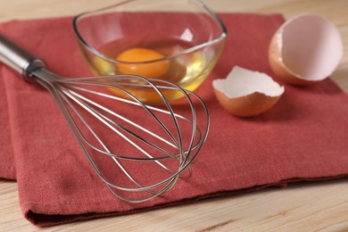 Metal whisk, raw eggs in bowl and shells on wooden table, closeup