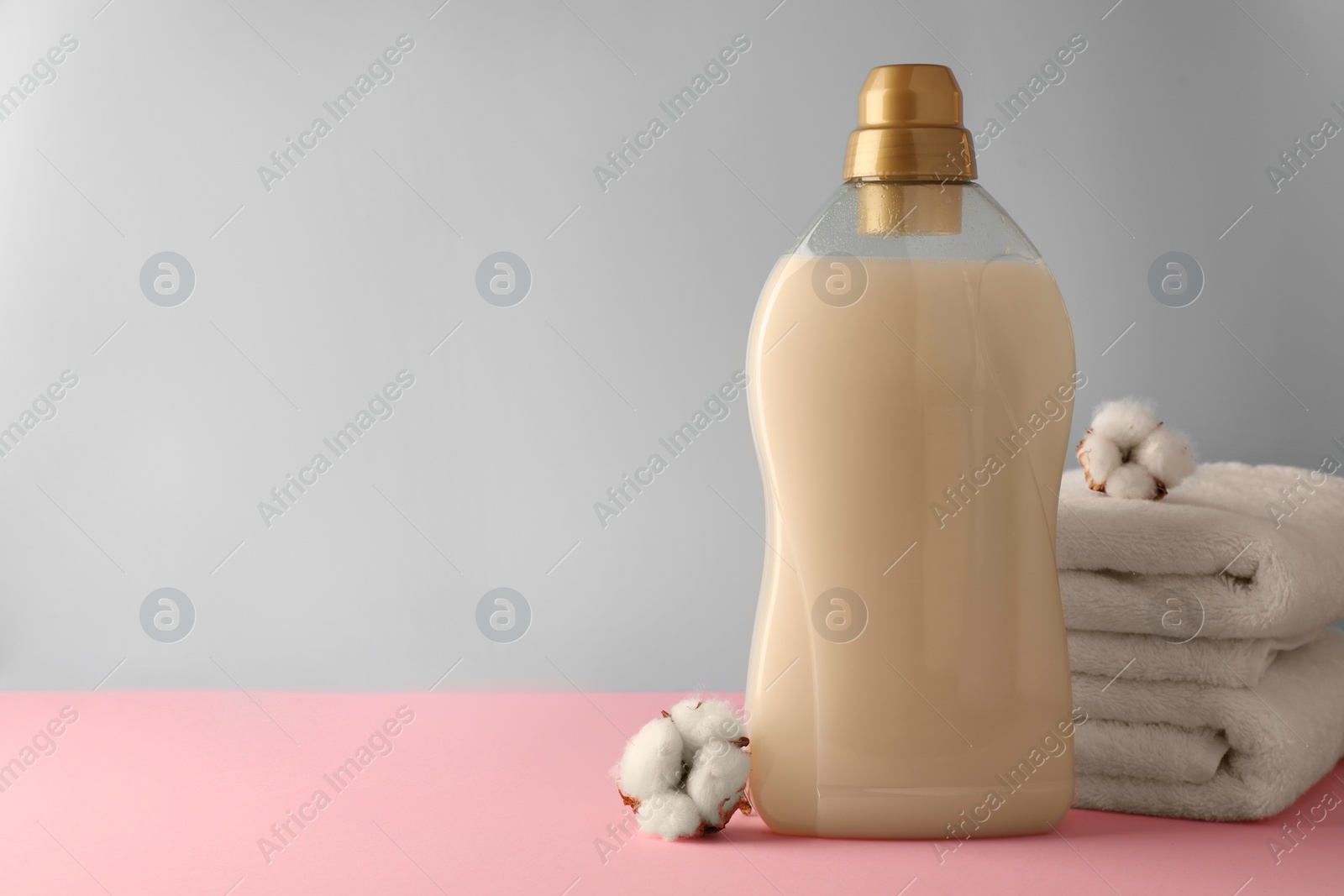 Photo of Bottle of fabric softener, stacked clean towels and fluffy cotton flowers on pink table against light grey background, space for text