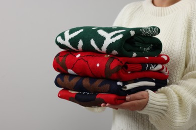Photo of Woman holding stack of different Christmas sweaters on light grey background, closeup
