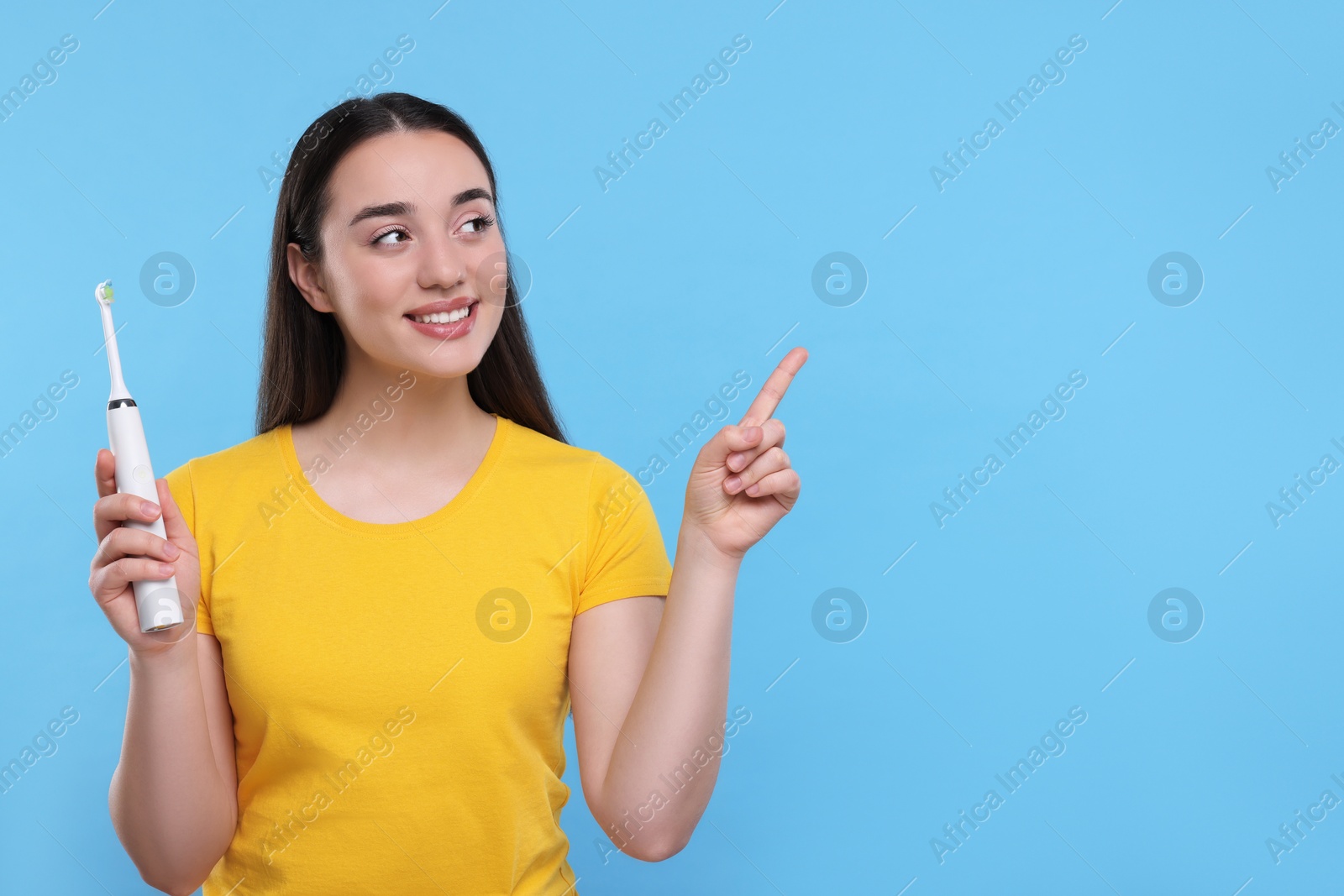 Photo of Happy young woman holding electric toothbrush and pointing on light blue background, space for text
