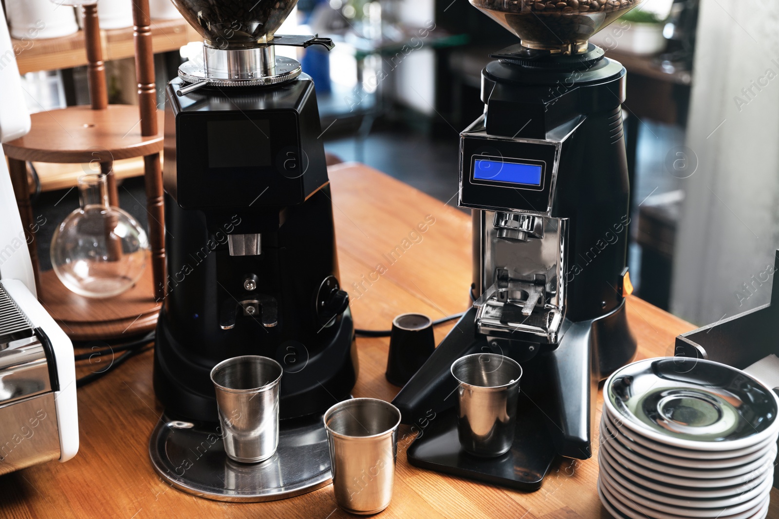 Photo of Modern coffee grinding machines with metal cups and saucers on wooden bar counter