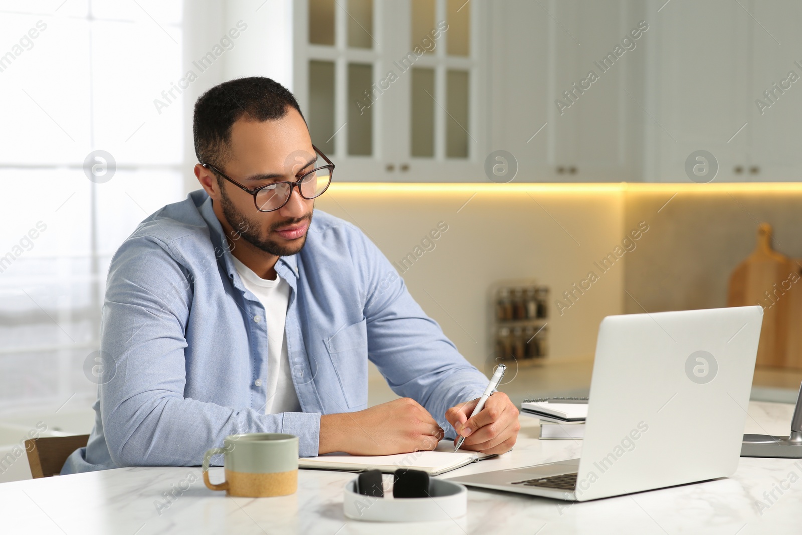 Photo of Young man with laptop writing in notebook at desk in kitchen. Home office