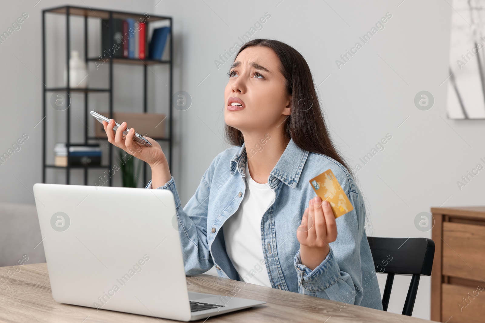 Photo of Confused woman with credit card, smartphone and laptop at table indoors. Be careful - fraud