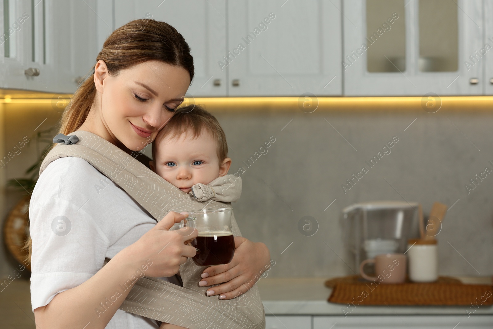 Photo of Mother with cup of drink holding her child in sling (baby carrier) in kitchen