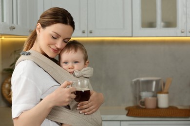 Photo of Mother with cup of drink holding her child in sling (baby carrier) in kitchen