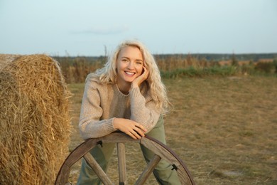 Photo of Beautiful woman posing with wooden cart wheel near hay bale in field. Autumn season