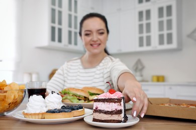 Happy overweight woman at table with unhealthy food in kitchen, focus on cake
