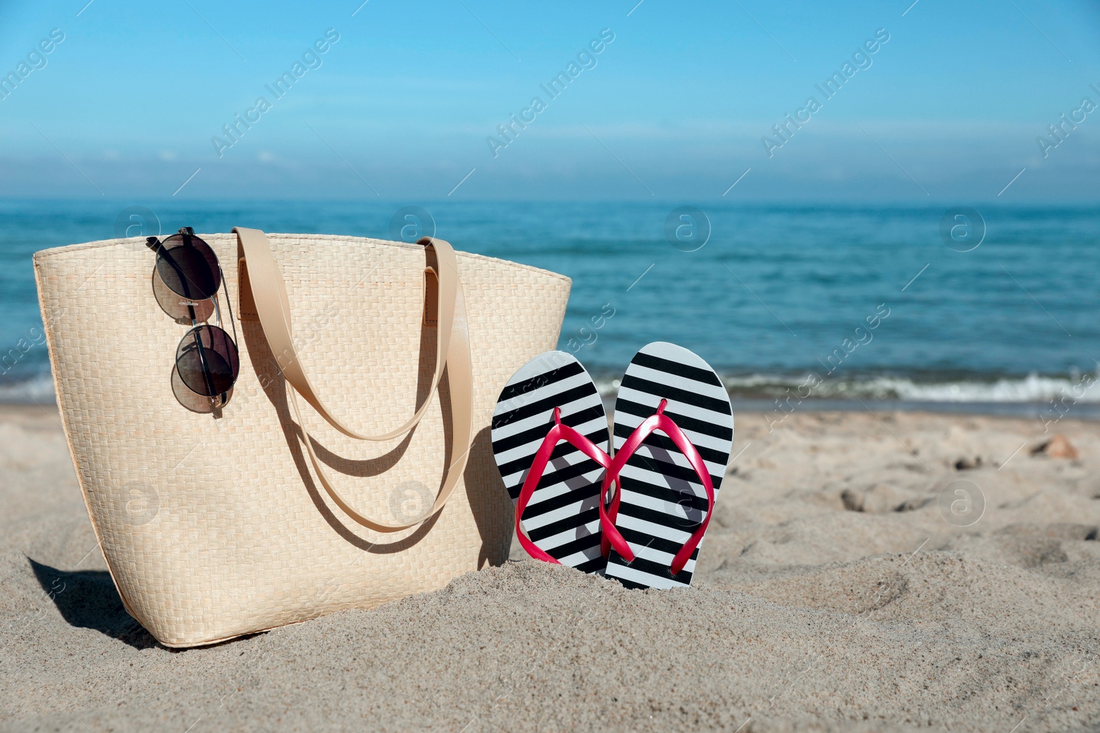Photo of Striped flip flops, sunglasses and beach bag on sandy seashore, space for text