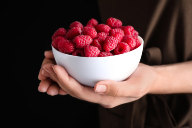 Photo of Woman holding bowl of delicious ripe raspberries on black background, closeup