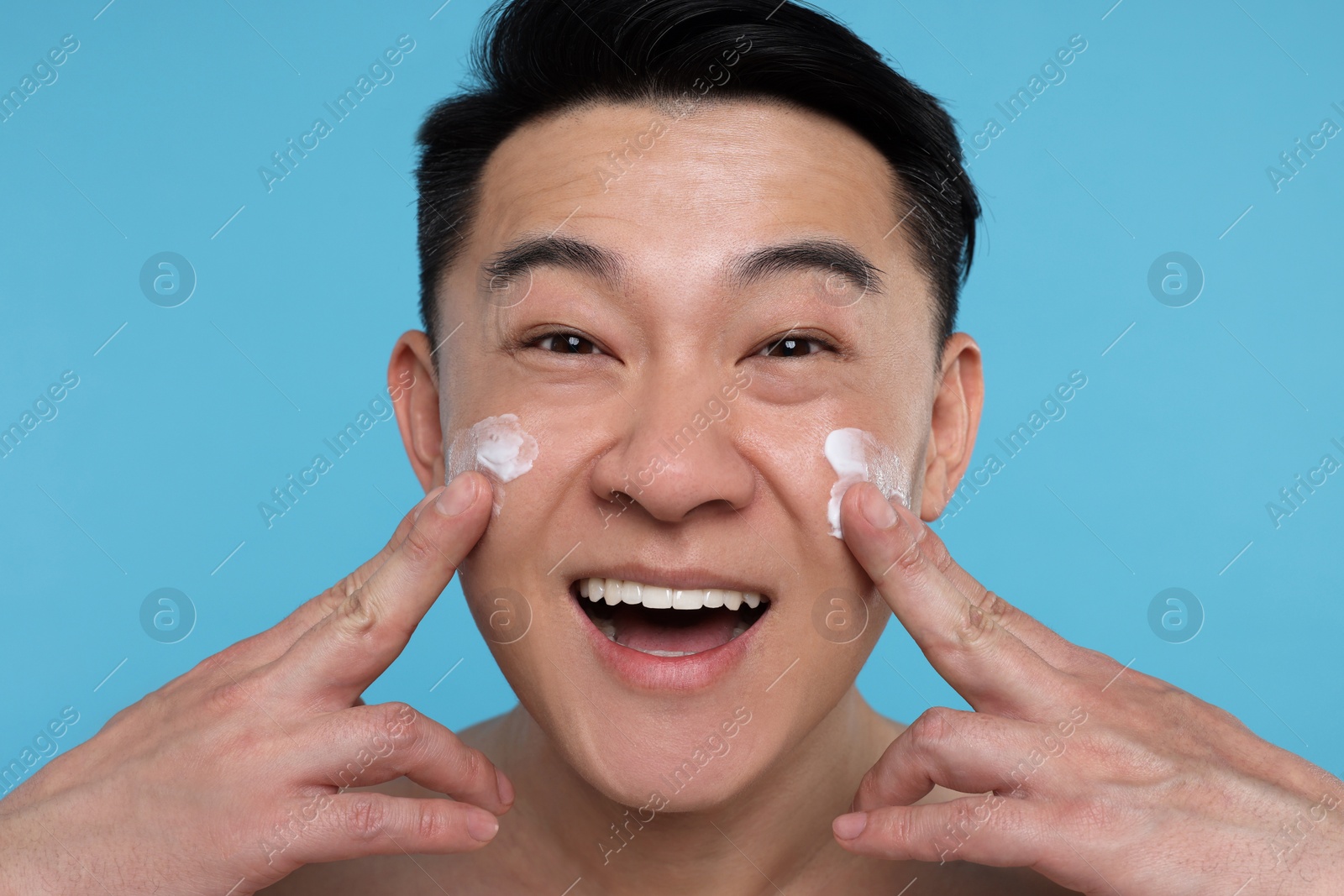 Photo of Happy man applying cream onto his face on light blue background, closeup