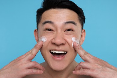 Happy man applying cream onto his face on light blue background, closeup