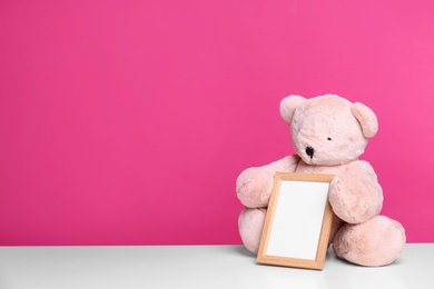 Photo of Photo frame and adorable teddy bear on table against color background, space for text. Child room elements