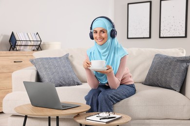 Muslim woman with cup of drink using laptop at wooden table in room