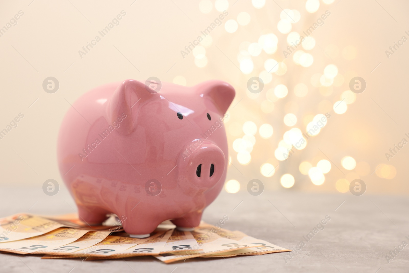 Photo of Piggy bank with euro banknotes on grey table against blurred lights, space for text