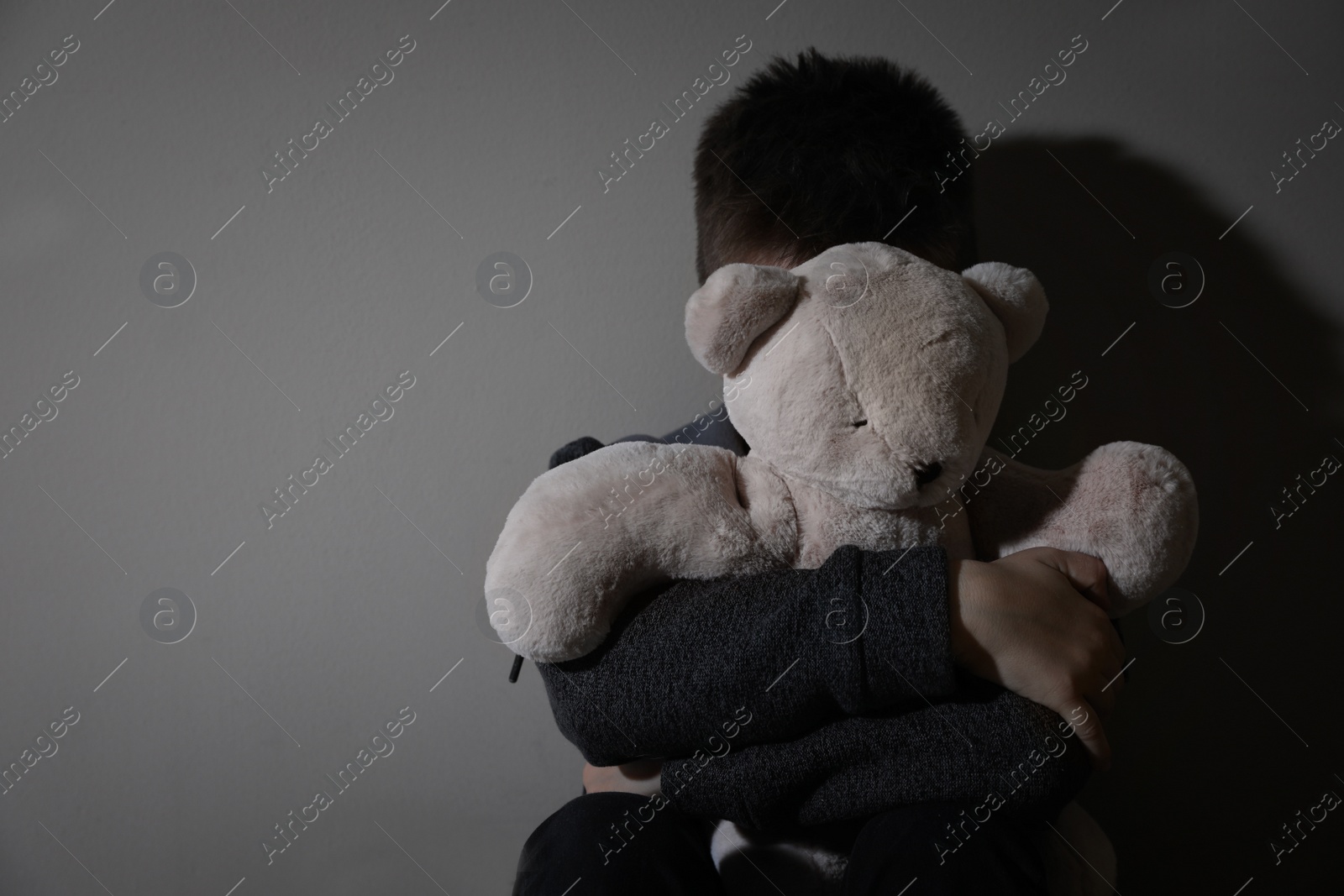 Photo of Scared little boy with teddy bear near beige wall, space for text. Domestic violence concept