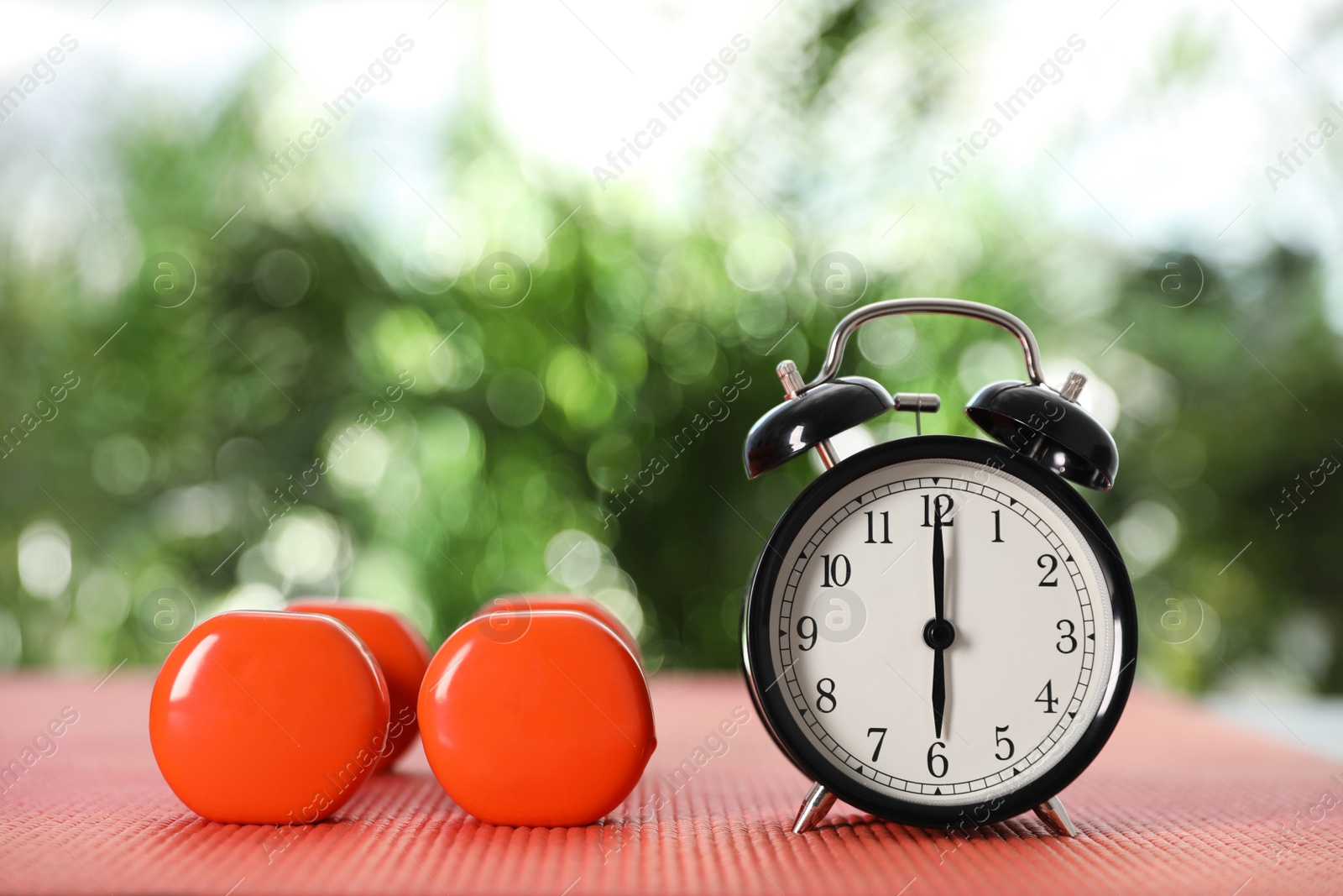 Photo of Alarm clock and dumbbells on yoga mat against blurred background. Morning exercise